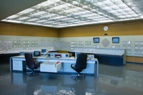 A control room with a large panel of monitors and controls, several office chairs, and desks with electronic equipment. The room is well-lit with a grid of fluorescent lights on the ceiling and has a clean, organized appearance.