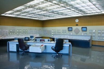 A control room with a large panel of monitors and controls, several office chairs, and desks with electronic equipment. The room is well-lit with a grid of fluorescent lights on the ceiling and has a clean, organized appearance.