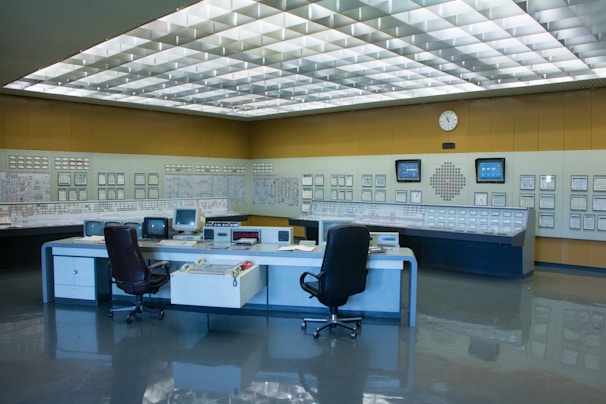A control room with a large panel of monitors and controls, several office chairs, and desks with electronic equipment. The room is well-lit with a grid of fluorescent lights on the ceiling and has a clean, organized appearance.