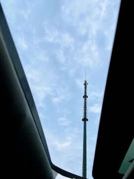 A tall communication tower stands against a partly cloudy sky, viewed through a car sunroof. The tower is mostly metallic and cylindrical with various antennae attached.