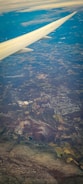 The image captures an aerial view of a landscape seen from an airplane. A wing is prominently visible extending from the top-left corner. Below, a patchwork of fields, forests, and towns stretches across the terrain. Clouds are scattered lightly across the sky, with a mixture of clear and cloudy weather visible.