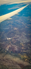 The image captures an aerial view of a landscape seen from an airplane. A wing is prominently visible extending from the top-left corner. Below, a patchwork of fields, forests, and towns stretches across the terrain. Clouds are scattered lightly across the sky, with a mixture of clear and cloudy weather visible.