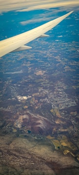 The image captures an aerial view of a landscape seen from an airplane. A wing is prominently visible extending from the top-left corner. Below, a patchwork of fields, forests, and towns stretches across the terrain. Clouds are scattered lightly across the sky, with a mixture of clear and cloudy weather visible.