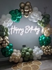 A decorative arrangement featuring a large arch made of balloons in gold, green, and white. A neon sign in white light spells out 'Happy Birthday' in the center. The floor is decorated with round woven mats adding a rustic touch.