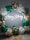 A decorative arrangement featuring a large arch made of balloons in gold, green, and white. A neon sign in white light spells out 'Happy Birthday' in the center. The floor is decorated with round woven mats adding a rustic touch.