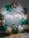 A decorative arrangement featuring a large arch made of balloons in gold, green, and white. A neon sign in white light spells out 'Happy Birthday' in the center. The floor is decorated with round woven mats adding a rustic touch.