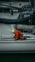 A person in a bright orange and yellow rain suit is kneeling on a white sailboat at a marina, surrounded by other boats and dock lines. The person appears to be adjusting or checking something on the deck.
