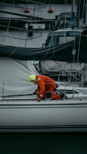 A person in a bright orange and yellow rain suit is kneeling on a white sailboat at a marina, surrounded by other boats and dock lines. The person appears to be adjusting or checking something on the deck.