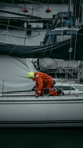 A person in a bright orange and yellow rain suit is kneeling on a white sailboat at a marina, surrounded by other boats and dock lines. The person appears to be adjusting or checking something on the deck.