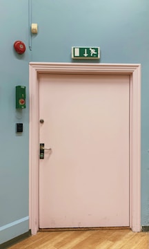 A light pink door set in a light blue wall, featuring an emergency exit sign with a running figure and a down arrow above the door. To the left of the door, there is a red alarm bell and a green emergency system box mounted on the wall. The floor is wooden.