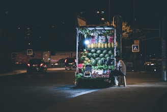 A night scene featuring a small, well-lit street vendor selling fruits. The vendor is seated on a stool beside the stand, which prominently displays an assortment of fresh fruits including watermelons, melons, and other produce. The background reveals a dimly lit street with a few cars passing by or parked and some pedestrian crossing signs.