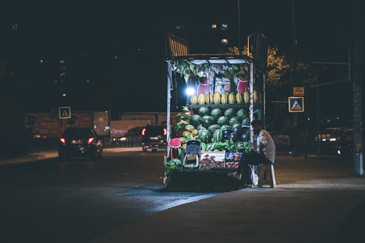 A night scene featuring a small, well-lit street vendor selling fruits. The vendor is seated on a stool beside the stand, which prominently displays an assortment of fresh fruits including watermelons, melons, and other produce. The background reveals a dimly lit street with a few cars passing by or parked and some pedestrian crossing signs.