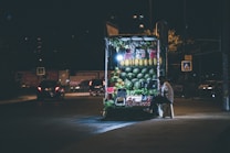 A night scene featuring a small, well-lit street vendor selling fruits. The vendor is seated on a stool beside the stand, which prominently displays an assortment of fresh fruits including watermelons, melons, and other produce. The background reveals a dimly lit street with a few cars passing by or parked and some pedestrian crossing signs.