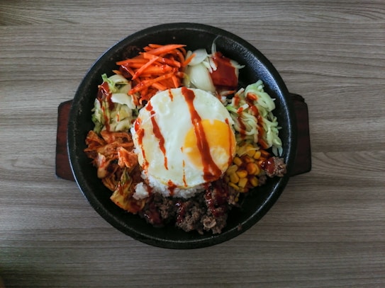 A sizzling black circular container holds a variety of food ingredients. At the center is a fried egg with a runny yolk, drizzled with a reddish-brown sauce. Surrounding the egg, there are colorful vegetables including shredded carrots, cabbage, onions, kimchi, corn, and chopped meat, all neatly arranged. The container is placed on a wooden surface.