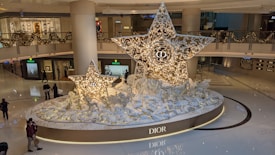 A large indoor display featuring two intricately designed, illuminated stars adorned with the Dior logo. Surrounding the stars are decorative white and gold elements, including reindeer figures and foliage. The setting is inside a shopping mall, with high-end store fronts such as Rolex visible. Holiday garlands decorate the mall's upper level.