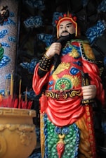 A colorful and intricately detailed statue of an ancient warrior, adorned in elaborate armor with vibrant reds, blues, and golds. The figure holds a staff and stands amidst an ornate backdrop featuring swirling clouds and elaborate patterns. In the foreground, a pot filled with incense sticks hints at a ritual or spiritual setting.
