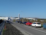 A road lined with parked cars extends toward a large industrial building in the distance. The building is surrounded by trees and features a prominent silvery tower. Overhead streetlights are spaced along the road, and the sky is clear and blue.