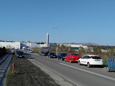A road lined with parked cars extends toward a large industrial building in the distance. The building is surrounded by trees and features a prominent silvery tower. Overhead streetlights are spaced along the road, and the sky is clear and blue.