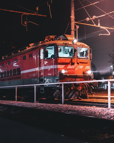 A red electric train is stopped on the railway tracks at night. The scene is illuminated by artificial lighting, highlighting the front and the side of the train. Overhead wires and a metal post for electric power supply are visible against the dark sky. The foreground includes a metal railing and gravel-covered ground.