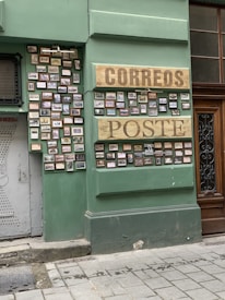 A green wall with numerous small frames displaying pictures. Two large wooden signs with the words 'CORREOS' and 'POSTE' are prominently featured. The frames are arranged neatly in rows. The setting appears to be part of a building exterior with an adjacent wooden door and a gray metal door on the left.