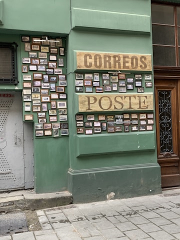 A green wall with numerous small frames displaying pictures. Two large wooden signs with the words 'CORREOS' and 'POSTE' are prominently featured. The frames are arranged neatly in rows. The setting appears to be part of a building exterior with an adjacent wooden door and a gray metal door on the left.