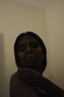 A person with dark hair and a septum piercing is adorned with golden glitter on their face and lips, creating a shimmering effect. The lighting is dim, casting shadows and giving an intimate, moody atmosphere. The person has their eyes closed and their hand is gently touching their shoulder, suggesting a sense of calm or introspection.