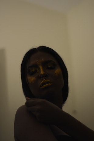 A person with dark hair and a septum piercing is adorned with golden glitter on their face and lips, creating a shimmering effect. The lighting is dim, casting shadows and giving an intimate, moody atmosphere. The person has their eyes closed and their hand is gently touching their shoulder, suggesting a sense of calm or introspection.