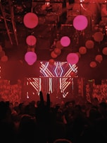 A nightclub scene with dim lighting and a red color scheme. Hanging spherical lights fill the ceiling, casting a warm glow. A crowd of people is gathered, with a large LED screen displaying abstract patterns at the front. The scene is dynamic and lively, suggestive of a party or concert.