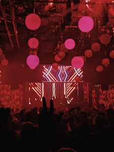 A nightclub scene with dim lighting and a red color scheme. Hanging spherical lights fill the ceiling, casting a warm glow. A crowd of people is gathered, with a large LED screen displaying abstract patterns at the front. The scene is dynamic and lively, suggestive of a party or concert.