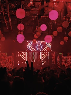 A nightclub scene with dim lighting and a red color scheme. Hanging spherical lights fill the ceiling, casting a warm glow. A crowd of people is gathered, with a large LED screen displaying abstract patterns at the front. The scene is dynamic and lively, suggestive of a party or concert.