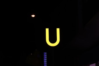 A bright yellow letter 'U' is illuminated against a dark background, likely representing a subway or metro station sign. A small, dimly lit streetlight and part of another building with illuminated text can be seen in the background.