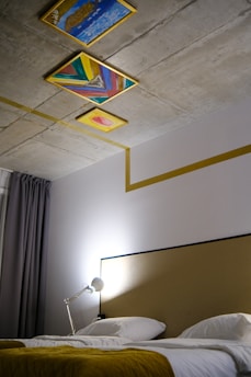 A modern bedroom features a bed with white linens and a mustard-colored throw. The headboard is simple, and a silver desk lamp is positioned on a side table. The ceiling is decorated with three colorful, abstract paintings with a geometric, artistic design. Grey curtains hang beside the bed.
