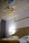 A modern bedroom features a bed with white linens and a mustard-colored throw. The headboard is simple, and a silver desk lamp is positioned on a side table. The ceiling is decorated with three colorful, abstract paintings with a geometric, artistic design. Grey curtains hang beside the bed.