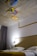 A modern bedroom features a bed with white linens and a mustard-colored throw. The headboard is simple, and a silver desk lamp is positioned on a side table. The ceiling is decorated with three colorful, abstract paintings with a geometric, artistic design. Grey curtains hang beside the bed.