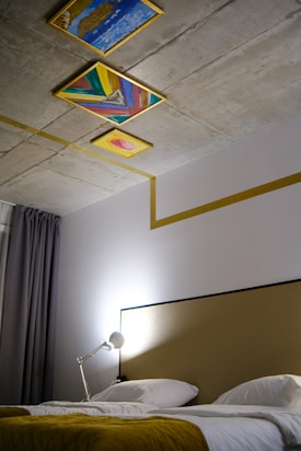 A modern bedroom features a bed with white linens and a mustard-colored throw. The headboard is simple, and a silver desk lamp is positioned on a side table. The ceiling is decorated with three colorful, abstract paintings with a geometric, artistic design. Grey curtains hang beside the bed.