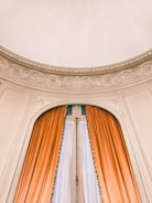 A grand arched window adorned with intricate plaster molding above, featuring long, elegant orange curtains framing it. The delicate details in the plasterwork add an element of sophistication.