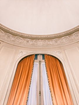 A grand arched window adorned with intricate plaster molding above, featuring long, elegant orange curtains framing it. The delicate details in the plasterwork add an element of sophistication.
