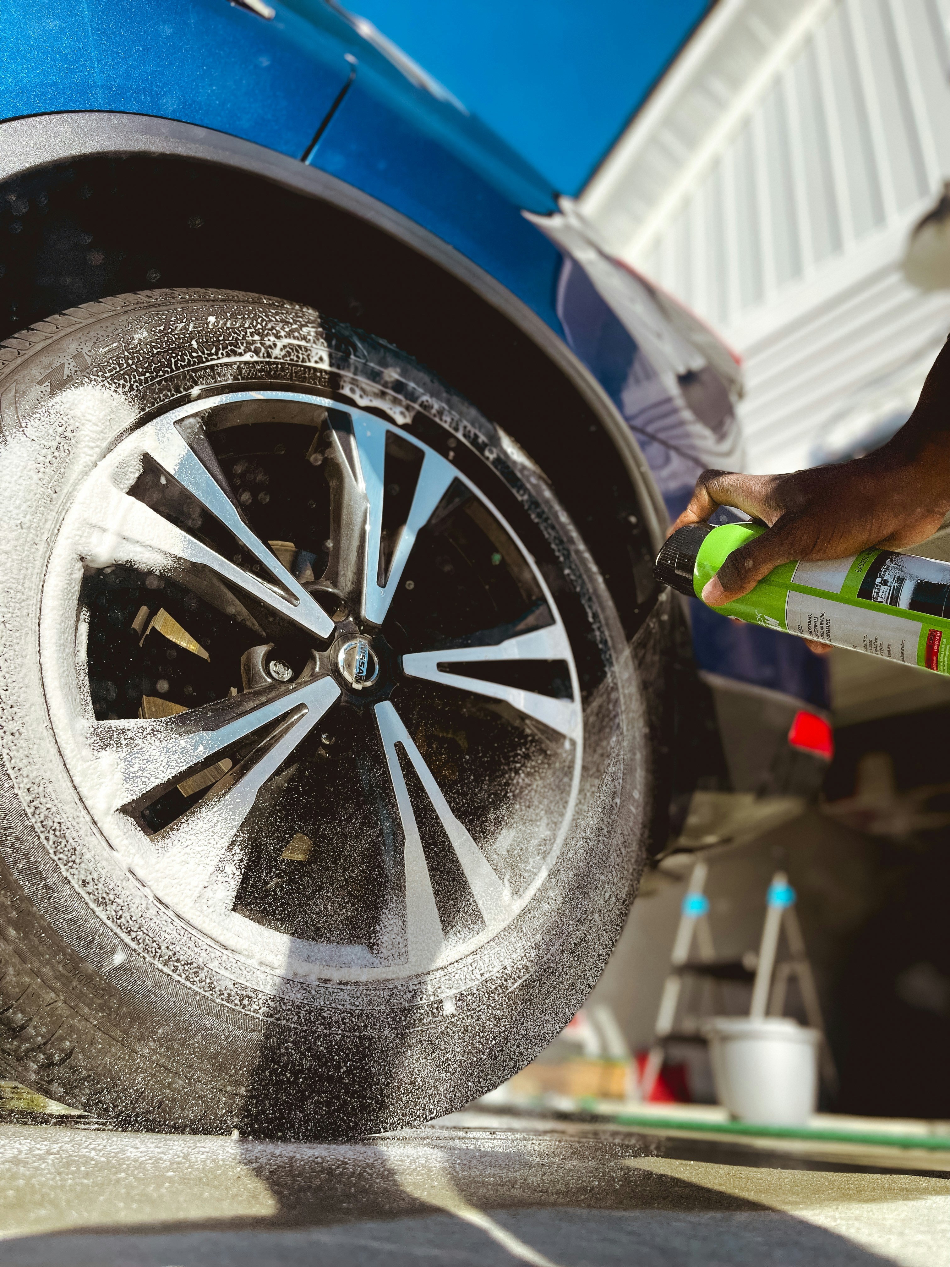 A close-up view of a car wheel being sprayed with a cleaning product. Foam is visible on the tire, and a hand is holding a green spray can. The car is a dark blue color and there is a clear sky in the background. The side of a building and a ladder are also visible in the scene.