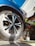 A close-up view of a car wheel being sprayed with a cleaning product. Foam is visible on the tire, and a hand is holding a green spray can. The car is a dark blue color and there is a clear sky in the background. The side of a building and a ladder are also visible in the scene.