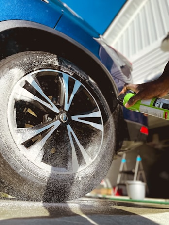 A close-up view of a car wheel being sprayed with a cleaning product. Foam is visible on the tire, and a hand is holding a green spray can. The car is a dark blue color and there is a clear sky in the background. The side of a building and a ladder are also visible in the scene.
