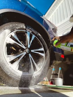A close-up view of a car wheel being sprayed with a cleaning product. Foam is visible on the tire, and a hand is holding a green spray can. The car is a dark blue color and there is a clear sky in the background. The side of a building and a ladder are also visible in the scene.