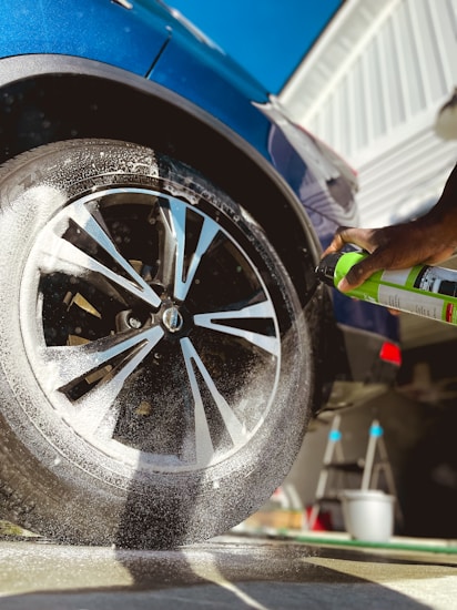 A close-up view of a car wheel being sprayed with a cleaning product. Foam is visible on the tire, and a hand is holding a green spray can. The car is a dark blue color and there is a clear sky in the background. The side of a building and a ladder are also visible in the scene.