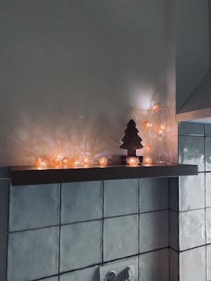 A wooden shelf is decorated with a string of orange fairy lights that cast a warm glow. The shelf holds a small wooden tree silhouette, a glass with some fairy lights inside, and a small wrapped box. The wall beneath is tiled, and there is a visible power outlet below the shelf.