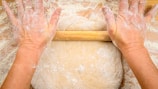 Hands rolling dough on a wooden board with flour scattered around.