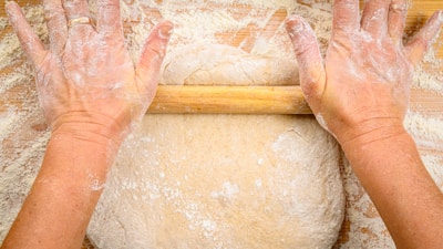 Hands rolling dough on a wooden board with flour scattered around.