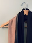 A wooden hanger is mounted on a light-colored wall. Draped over it are two pieces of fabric; one is beige with a soft texture and the other is black with a smooth, leather-like strip.