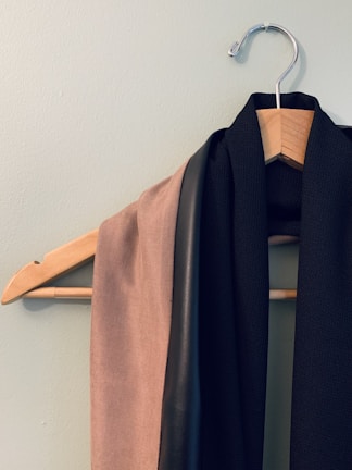 A wooden hanger is mounted on a light-colored wall. Draped over it are two pieces of fabric; one is beige with a soft texture and the other is black with a smooth, leather-like strip.
