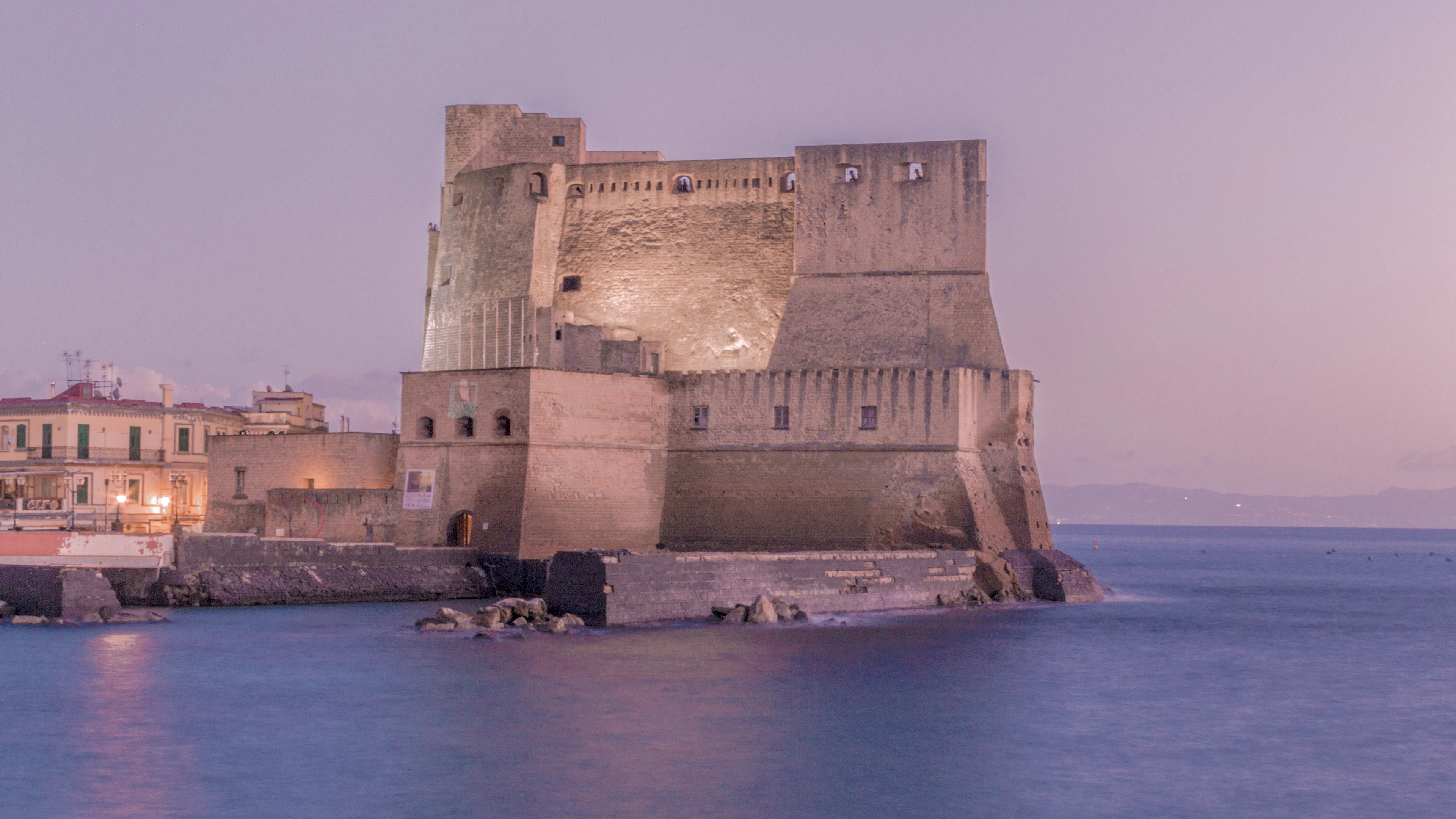 Castel dell'Ovo at dusk, its floodlit stone bulk rising straight out of a still lilac-pink sea, with the lit windows of the Santa Lucia waterfront to one side.
