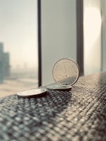 Several coins are placed on a textured surface near a window, capturing a bright, sunlit environment. The primary focus is on a coin from the United Arab Emirates, displaying Arabic script. The background features a blurred cityscape with tall buildings visible through the window.