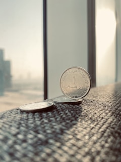 Several coins are placed on a textured surface near a window, capturing a bright, sunlit environment. The primary focus is on a coin from the United Arab Emirates, displaying Arabic script. The background features a blurred cityscape with tall buildings visible through the window.